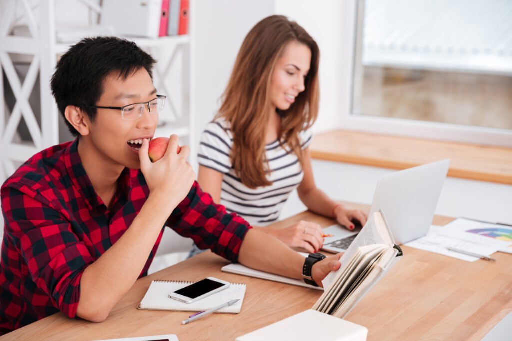 Asian student eating apple and reading while groupmate working at laptop