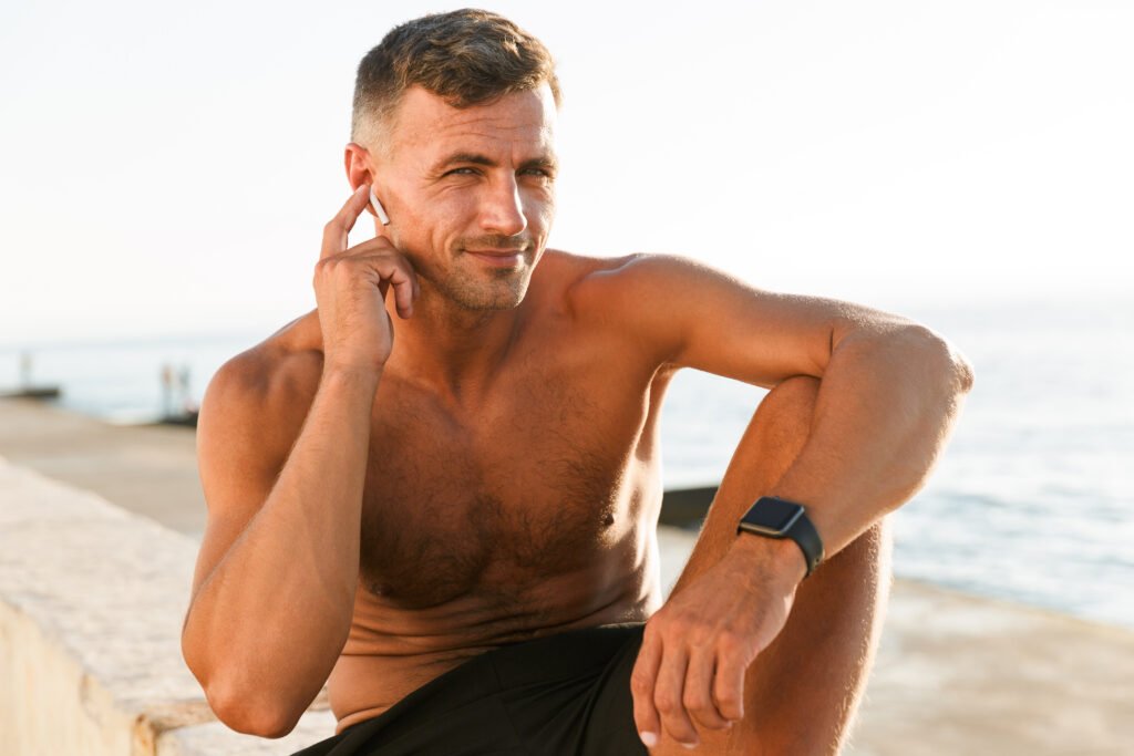 Happy handsome shirtless sportsman with earphones resting at the beach